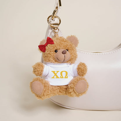The Darling Effect's Chi Omega Teddy Bear Bag Charm—a small brown bear with a red bow and white shirt with yellow ΧΩ letters—hangs from a white handbag, making it an ideal sorority gift or cute accessory.