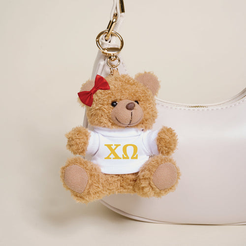 The Darling Effect's Chi Omega Teddy Bear Bag Charm—a small brown bear with a red bow and white shirt with yellow ΧΩ letters—hangs from a white handbag, making it an ideal sorority gift or cute accessory.