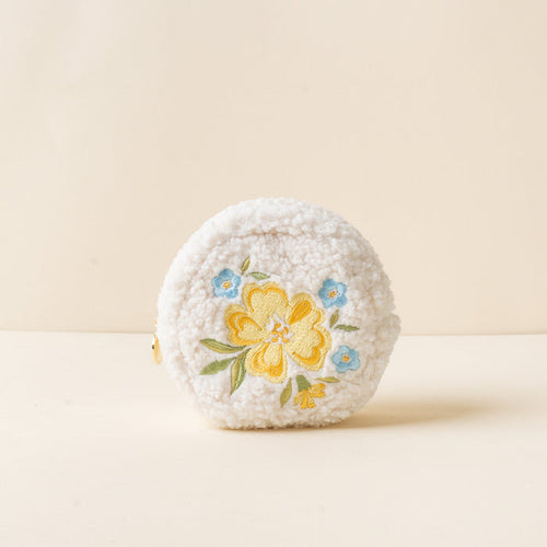 The-darling-effect’s Circle Dainty Floral Yellow Teddy Pouch is a small, round, white sherpa pouch with embroidered yellow and blue flowers on the front, shown on a neutral beige background.