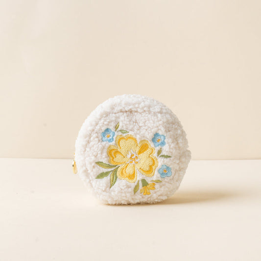 The-darling-effect’s Circle Dainty Floral Yellow Teddy Pouch is a small, round, white sherpa pouch with embroidered yellow and blue flowers on the front, shown on a neutral beige background.