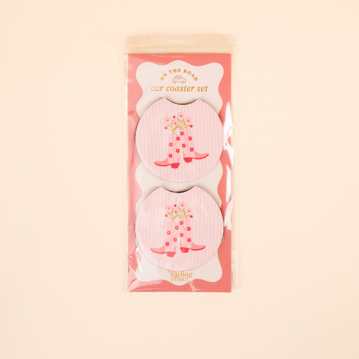 The Darling Effect’s Cowgirl Boots On the Road Car Coaster Set includes two round car coasters with a pink and white striped design, red cowboy boots, and flowers—perfect for adding style to your car cup holder.
