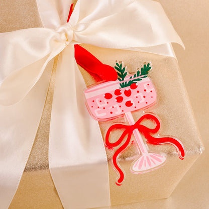 A gold-wrapped gift features a cream satin bow and The Darling Effect Cranberry Cosmo Holiday Tree Ornament—a pink festive drink with red cherries and a bow—perfect for holiday season and festive décor.