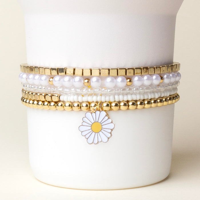 The Daisy Dream Tumbler Bracelet Stack by the-darling-effect features gold, white pearl, and beaded bracelets with a daisy flower charm, displayed on a white holder against a light background. Perfect for styling your tumbler!.