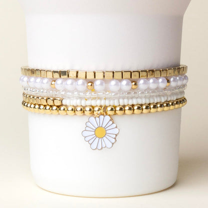 The Daisy Dream Tumbler Bracelet Stack by the-darling-effect features gold, white pearl, and beaded bracelets with a daisy flower charm, displayed on a white holder against a light background. Perfect for styling your tumbler!.