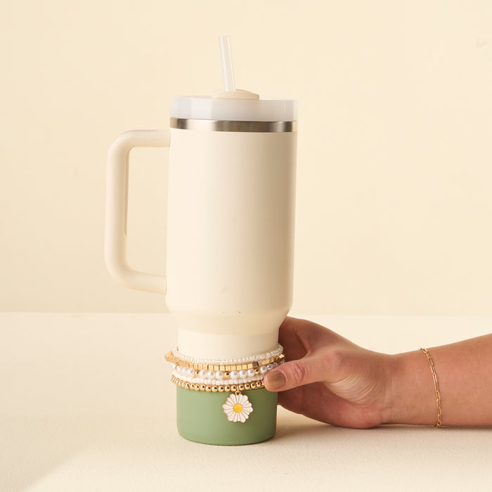 A hand holds a large cream and green 40 oz tumbler with a straw, adorned with the-darling-effect’s Daisy Dream Tumbler Bracelet Stack and a cute daisy sticker—fun inspiration for accessorizing your tumbler.