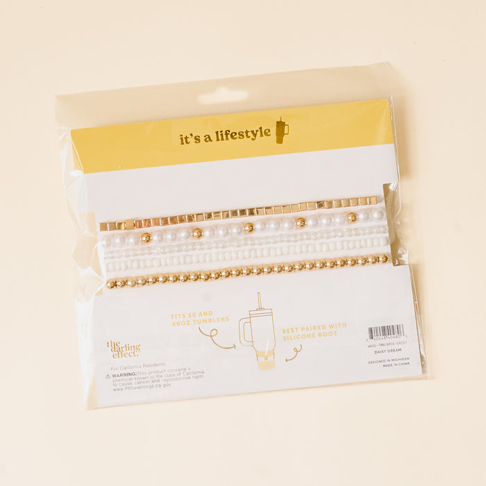 The Daisy Dream Tumbler Bracelet Stack by the-darling-effect features decorative gold, pearl, and white beaded bands for 30 & 40 oz tumblers. Labeled "it’s a lifestyle," these stylish accessories are shown on a beige background.