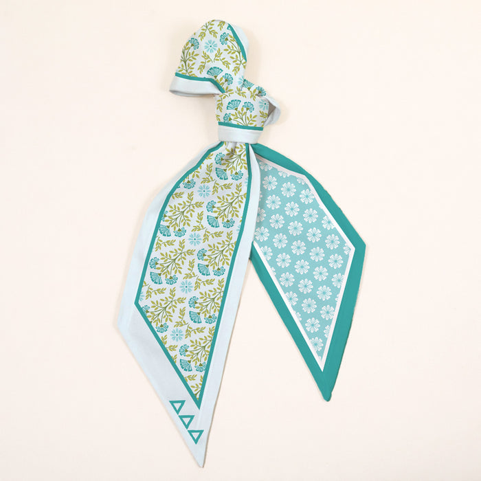 The Darling Effect’s Delta Delta Delta Twirl & Tie Scarf Accessory dazzles with a blue, green, and white floral print. Draped and tied with pointed ends, it features a geometric border on one side and a diamond floral motif on the other.