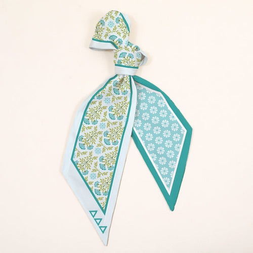 The Darling Effect’s Delta Delta Delta Twirl & Tie Scarf Accessory dazzles with a blue, green, and white floral print. Draped and tied with pointed ends, it features a geometric border on one side and a diamond floral motif on the other.