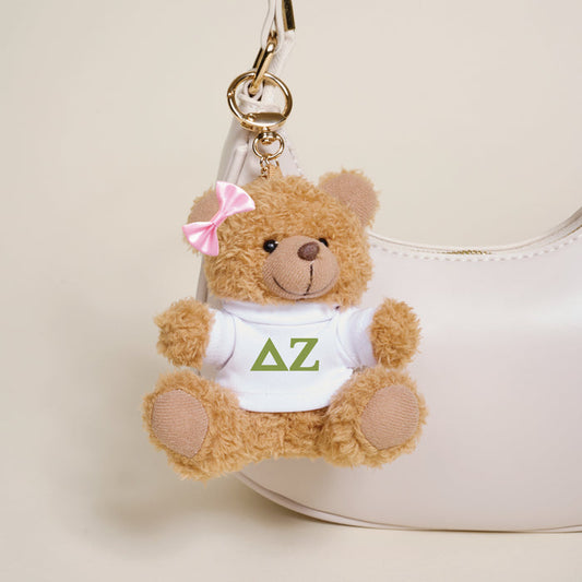 The Darling Effect's Delta Zeta Teddy Bear Bag Charm—a small brown bear with a pink bow and white shirt with green letters—is attached to a white handbag, making it a perfect accessory or Bid Day gift.