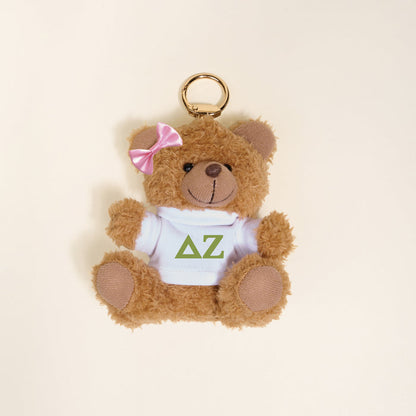 The Darling Effect’s Delta Zeta Teddy Bear Bag Charm features a small brown bear with a pink bow, wearing a white shirt with green ΔΖ letters—perfect as a Bid Day gift or cute accessory for your bag.