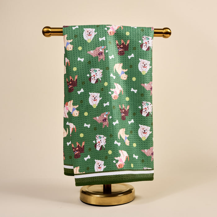 The Darling Effect's Dog Days Waffle Tea Towel, with cartoon dog faces, bones, and balls, is displayed on a gold stand against a beige background, bringing playful charm to any space.