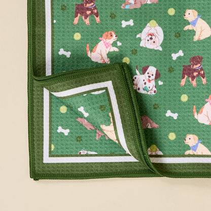 The Darling Effect’s Dog Days Waffle Tea Towel is green with a white border, showcasing playful dog, bone, and paw print illustrations. A folded corner reveals the same delightful pattern on the reverse side.