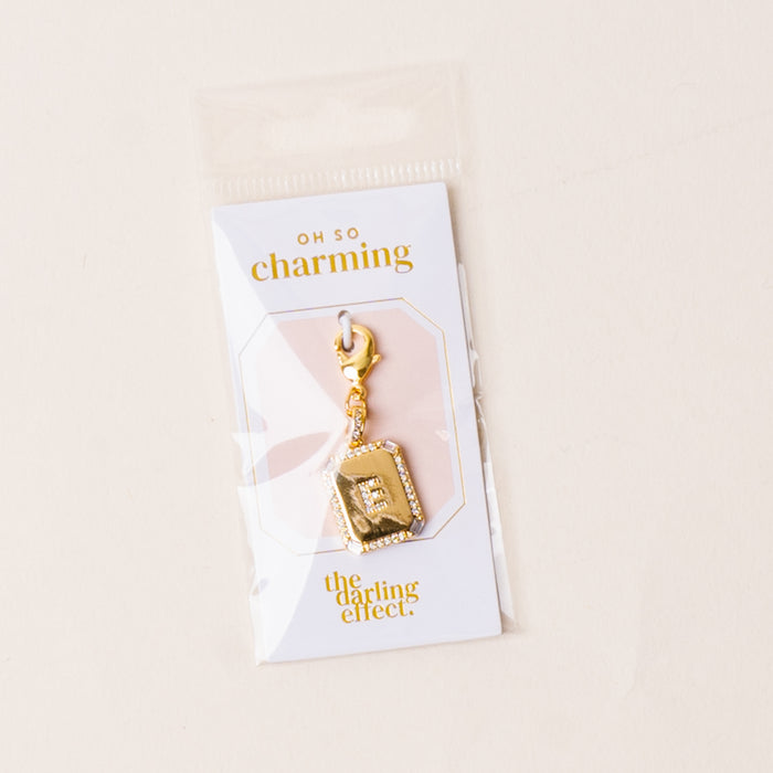 The-darling-effect’s E Jewelry Charm Initial Letter Gold features a gold-plated brass rectangular pendant engraved with “E,” attached to an Oh So Charming card and packaged in a clear plastic sleeve—ideal for personalized jewelry lovers.