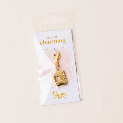 The-darling-effect’s E Jewelry Charm Initial Letter Gold features a gold-plated brass rectangular pendant engraved with “E,” attached to an Oh So Charming card and packaged in a clear plastic sleeve—ideal for personalized jewelry lovers.