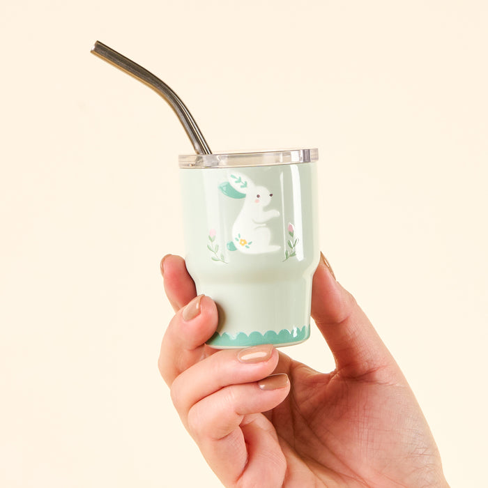 A hand holds the Easter Bunny 2 oz Tiny Tumbler by the-darling-effect. This light green mini cup features a white rabbit illustration surrounded by flowers and leaves, and comes with a metal straw.