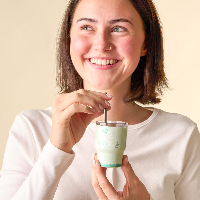 A young woman with short brown hair and a nose ring smiles while holding the-darling-effect Easter Bunny 2 oz Tiny Tumbler with a straw, wearing a white long-sleeve shirt against a light background.
