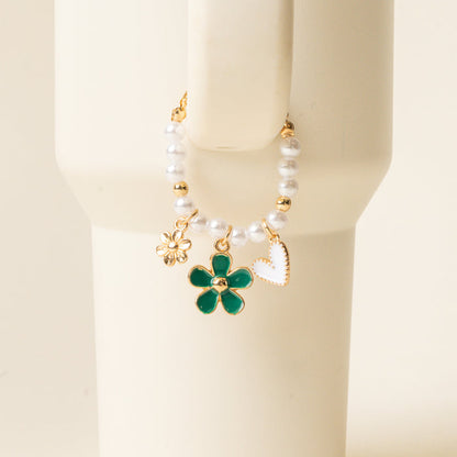 The-darling-effect Enchanted Tumbler Charm is a beaded bracelet with white pearls and three gold charms—flower, green enamel flower, and white heart—shown on a cream cylinder; ideal as an elegant tumbler accessory or paired with tumbler charms.