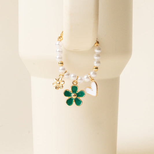 The-darling-effect Enchanted Tumbler Charm is a beaded bracelet with white pearls and three gold charms—flower, green enamel flower, and white heart—shown on a cream cylinder; ideal as an elegant tumbler accessory or paired with tumbler charms.