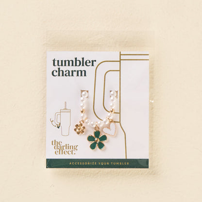 The Enchanted Tumbler Charm by the-darling-effect features a beaded chain with gold and green flower, gold flower, and pink heart charms. The packaging reads "Tumbler Charms" and "the darling effect." Accessorize your tumbler in style!.