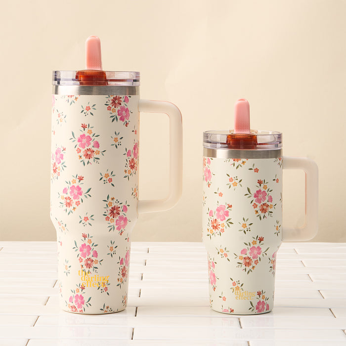 The-darling-effect’s Endless Daydream Cream 40 oz Lifestyle Flip Straw Tumbler and a matching floral tumbler with handles and pink straws sit side by side on a white surface against a beige background.