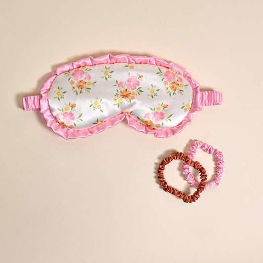 The Darling Effect’s Endless Daydream Cream Beauty Sleep Mask, featuring pink florals and a pink ruffled edge, rests beside brown and pink satin scrunchies on a beige background—a charming satin set for bedtime.