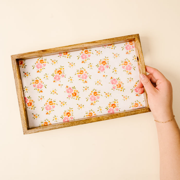 A hand holds the-darling-effect’s Endless Daydream Cream Rectangle Wood Enamel Tray, featuring a white background adorned with small pink, orange, yellow flowers, and green leaves—a charming piece for any table.