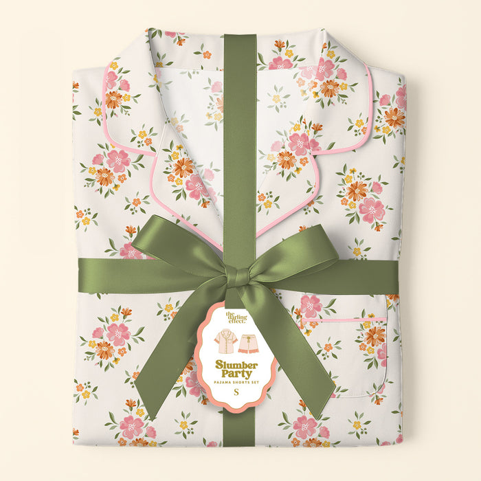 A folded Endless Daydream Cream Slumber Party Pajama Shorts Set by The Darling Effect, in ultra-soft fabric with pink trim and pink-orange flowers, is tied with a green ribbon and tagged for relaxed fit and dreamy comfort.