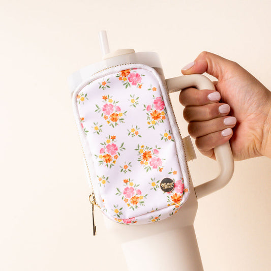 A hand holds the-darling-effect’s Endless Daydream Cream Tumbler Fanny Pack—a stylish cream water bottle with a handle and a white pouch featuring pink and orange floral prints. The person has light-colored nail polish.