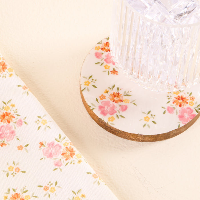 The Endless Daydream Cream Wood Enamel Coaster by the-darling-effect, with its pink and orange floral design, adds charm to any light surface, paired here with a clear glass and matching floral-patterned fabric.
