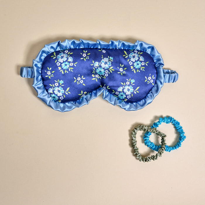 The Darling Effect Endless Daydream Navy Beauty Sleep Mask with a ruffled edge sits on beige beside teal and silver satin scrunchies—a charming trio to elevate your satin sleep set.