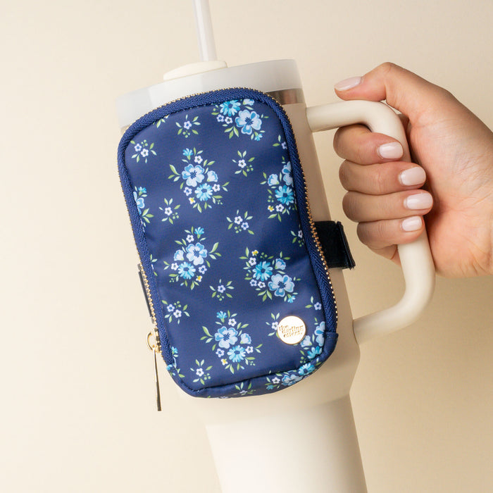 A hand holds a large white tumbler with a straw, featuring a stylish fanny pack-style navy blue floral zip pouch attached to the handle. The pouch has a gold zipper and a round gold logo.