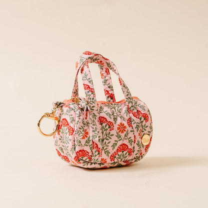 The Darling Effect’s Everbloom Dusty Rose Itty Bitty Duffle Bag Charm is a small, rounded handbag with dual handles, a red, orange, and green floral print on light background, gold keyring, quilted bag charm, and gold circular logo.