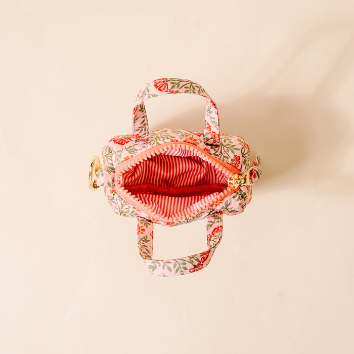 The Darling Effect’s Everbloom Dusty Rose Itty Bitty Duffle Bag Charm, featuring a floral pattern and two handles, is displayed from above on a light beige background. Its zippered top opens to reveal a red-and-white striped lining.