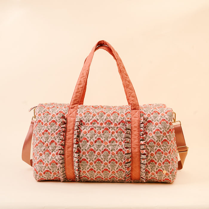 The Darling Effect’s Everbloom Dusty Rose On-the-Go Quilted Weekender Bag features a red and gray floral pattern, ruffled accents, two padded handles, a front pocket, and an adjustable shoulder strap on a pale beige background.