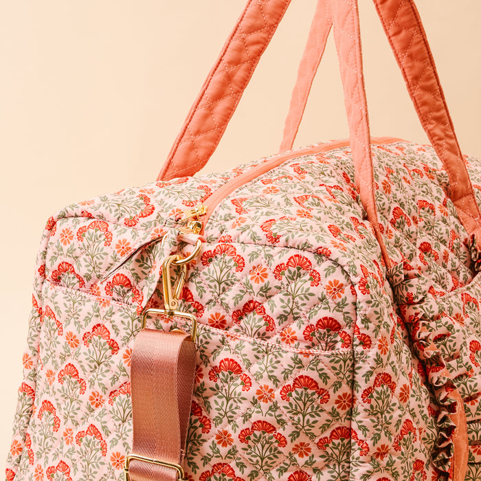 The Darling Effect Everbloom Dusty Rose On-the-Go Quilted Weekender Bag features pink handles, a floral red and green pattern, a gold zipper, and a detachable, adjustable pink shoulder strap.