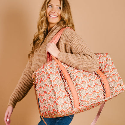 A smiling woman with long, wavy blonde hair in a tan sweater carries The Darling Effect’s Everbloom Dusty Rose On-the-Go Quilted Weekender Bag, featuring pink handles, over her shoulder against a beige background.