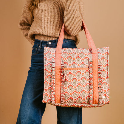 A woman in a brown sweater and blue jeans holds The Darling Effect’s Everbloom Dusty Rose The Everyday Quilted Tote Bag, featuring pink handles and floral designs, against a beige background.
