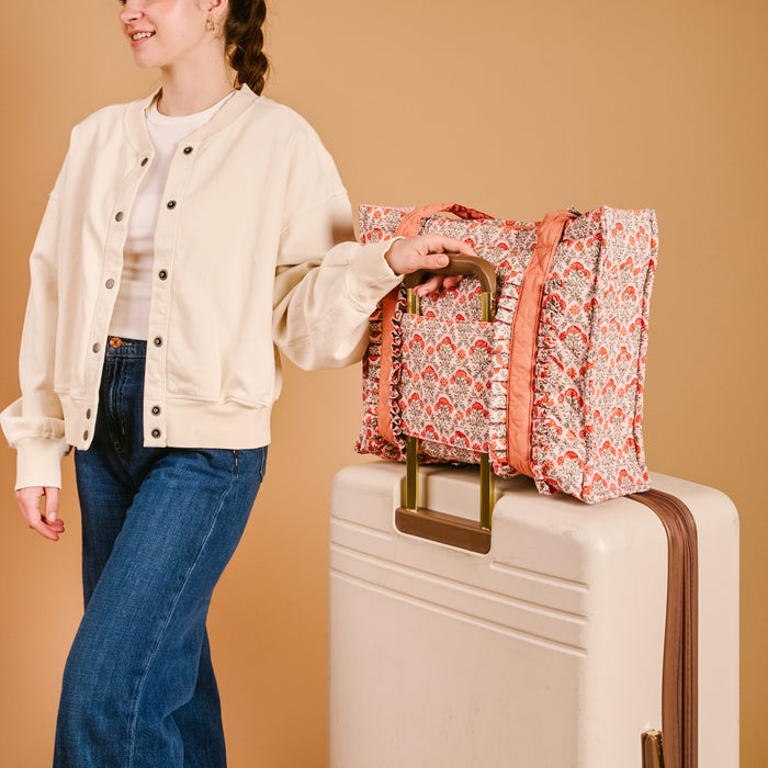 A woman in a cream jacket and blue jeans stands beside a large cream suitcase with The Darling Effect Everbloom Dusty Rose The Everyday Quilted Tote Bag resting on top, set against a solid light brown background.