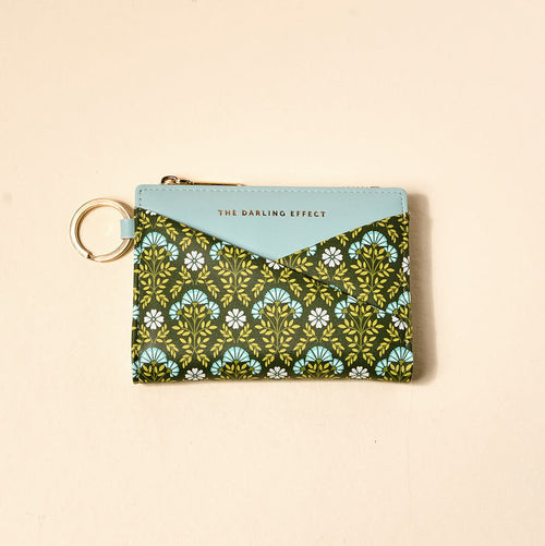 The Everbloom Forest Green Luxe Pocket Wallet by The Darling Effect is a compact vegan leather pouch with a criss-cross front, blue flap labeled with the brand, keyring, and green, blue, and white floral patterns on dark.