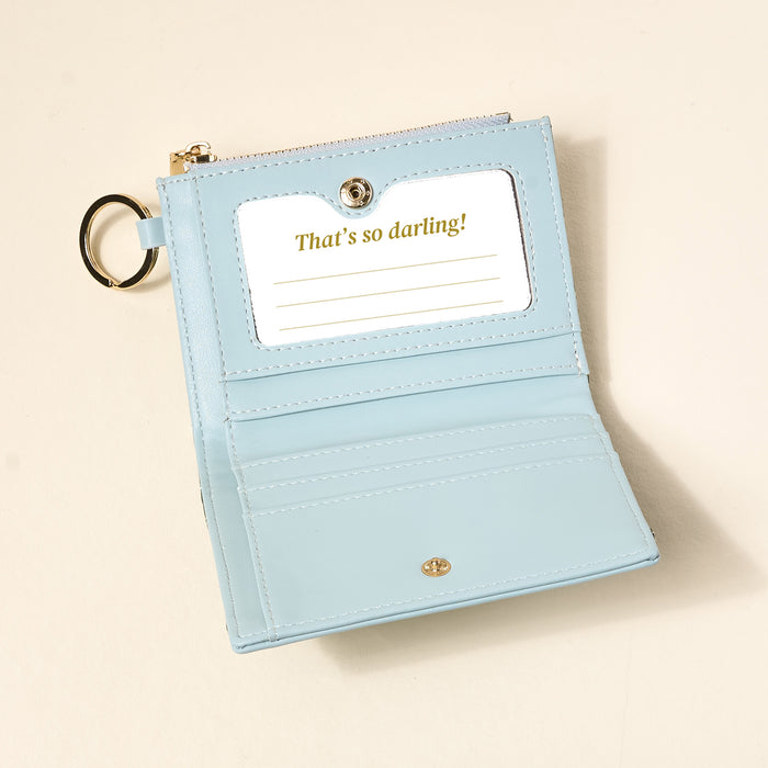 The Everbloom Forest Green Luxe Pocket Wallet by The Darling Effect is open, displaying card slots and a clear ID window with "Thats so darling!" text. A gold keychain ring is attached to the side.