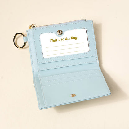 The Everbloom Forest Green Luxe Pocket Wallet by The Darling Effect is open, displaying card slots and a clear ID window with "Thats so darling!" text. A gold keychain ring is attached to the side.