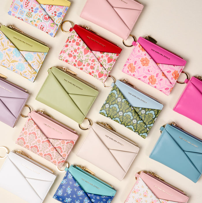 The Darling Effect's Everbloom Forest Green Luxe Pocket Wallet is displayed with other compact vegan leather wallets in pastel and floral patterns. Most feature envelope flaps, gold zippers, or criss-cross fronts on a light surface.