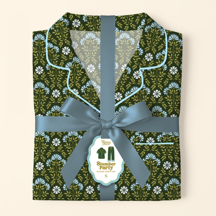 A folded Everbloom Forest Green Slumber Party Pajama Pants Set by The Darling Effect, in soft fabric with a floral print, is tied with a blue ribbon and bow. A tag shows the product name and a pajamas illustration.