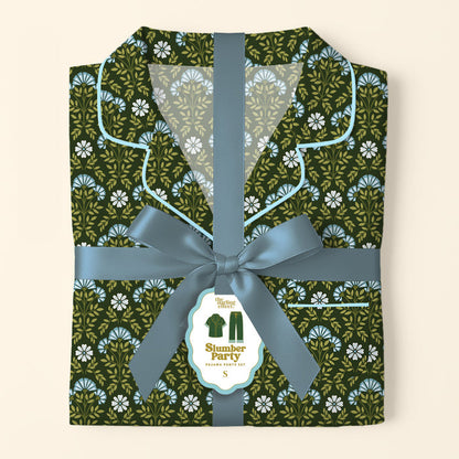 A folded Everbloom Forest Green Slumber Party Pajama Pants Set by The Darling Effect, in soft fabric with a floral print, is tied with a blue ribbon and bow. A tag shows the product name and a pajamas illustration.