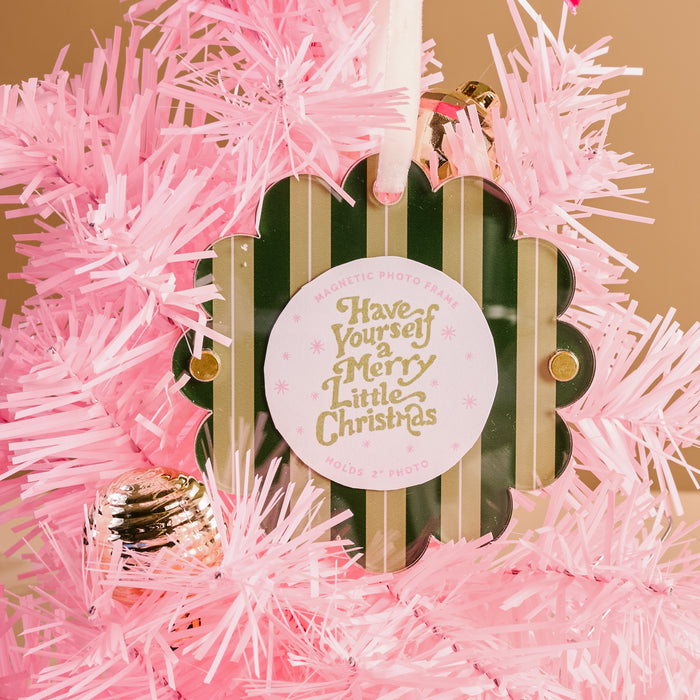 The Darling Effect's Evergreen Stripes Scalloped Round Picture Frame Ornament hangs on a pink artificial Christmas tree, featuring green and gold accents and the phrase “Have Yourself a Merry Little Christmas,” with a pink ornament nearby.
