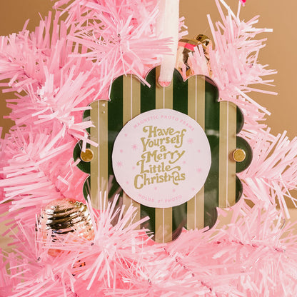 The Darling Effect's Evergreen Stripes Scalloped Round Picture Frame Ornament hangs on a pink artificial Christmas tree, featuring green and gold accents and the phrase “Have Yourself a Merry Little Christmas,” with a pink ornament nearby.