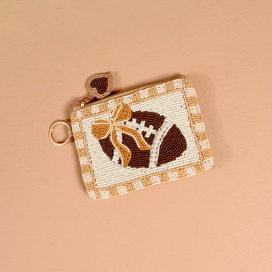 Introducing the Fab Football Handcrafted Beaded Pouch by The Darling Effect—a charming small pouch featuring a football design, embellished with a gold bow and finished with a heart-shaped zipper pull, displayed against a beige background.