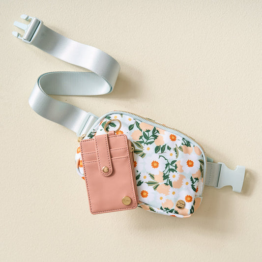 The Darling Effect's Flower Talk Print Belt Bag & Wallet Set features a floral-patterned bag with a light blue strap and buckle, plus a pink keychain wallet attached to the zipper pull, displayed on a beige surface.