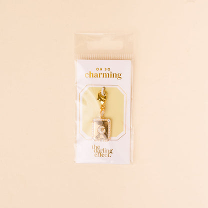 The-darling-effect’s G Jewelry Charm Initial Letter Gold features a gold-plated brass charm with rhinestones and a rectangular frame, hanging from a lobster clasp on an “Oh So Charming” card—perfect for lovers of personalized jewelry.