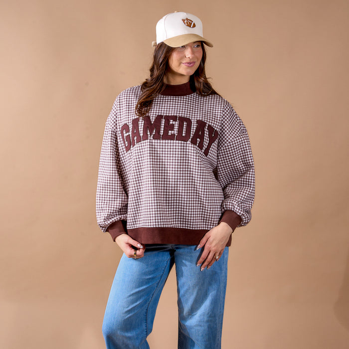 A woman, smiling gently, stands against a beige background wearing The Darling Effect’s Gameday Mockneck Sweatshirt in maroon and white checkered print, paired with blue jeans and a cream and brown cap.